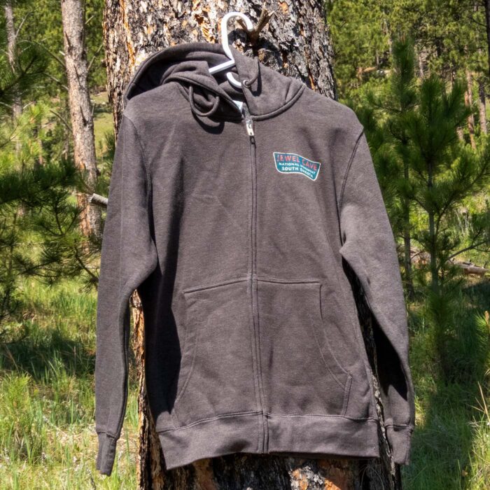 Charcoal Jewel Cave National Monument full-zip hoodie on a hanger in a Black Hills ponderosa pine forest, front