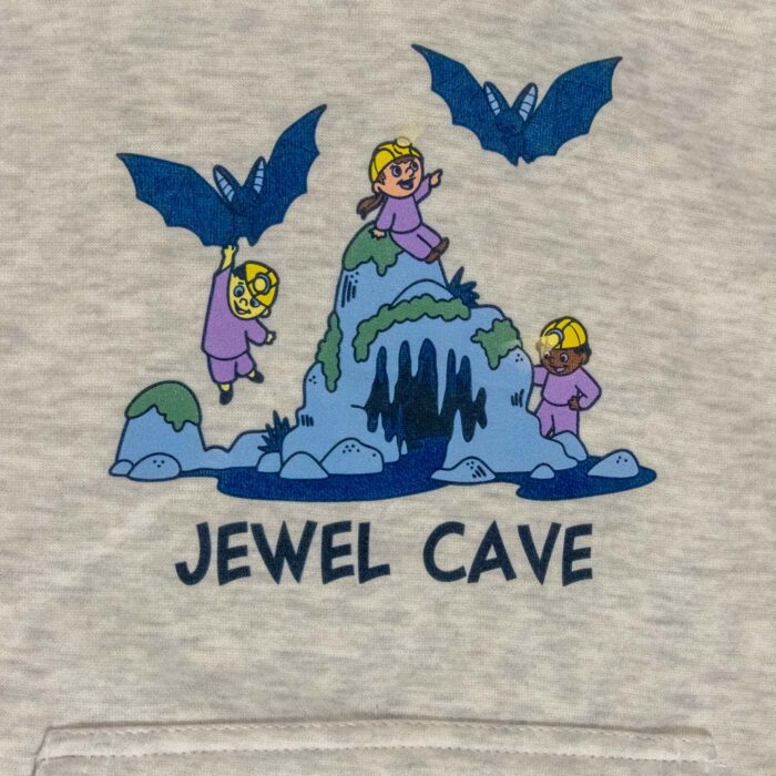 Close-up of the Jewel Cave youth hoodie front graphic: three kids in hard-hat headlamps climbing a blue cave rock with two bats overhead and Jewel Cave lettered below