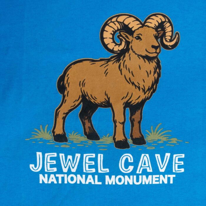 Close-up of the front print: a brown bighorn ram with full-curl cream horns standing in grass, above Jewel Cave in outlined white letters and National Monument in solid white
