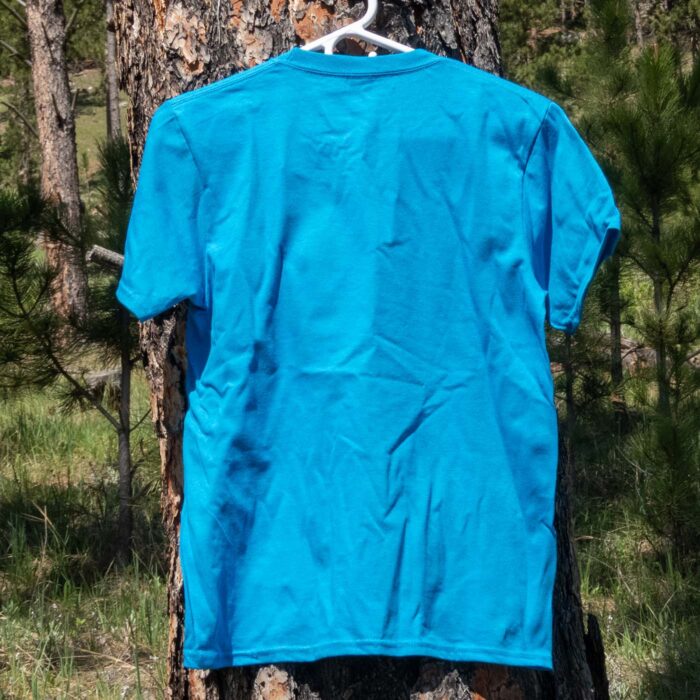 Bright blue Jewel Cave youth bighorn t-shirt on a hanger in a ponderosa pine forest, back view