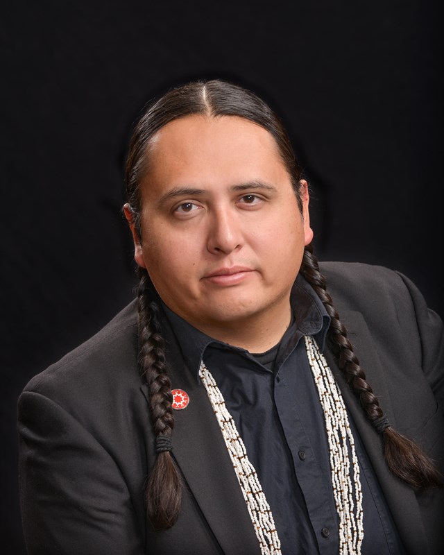 Portrait of artist Marty Two Bulls Jr., who has long braided hair and wears a beaded necklace