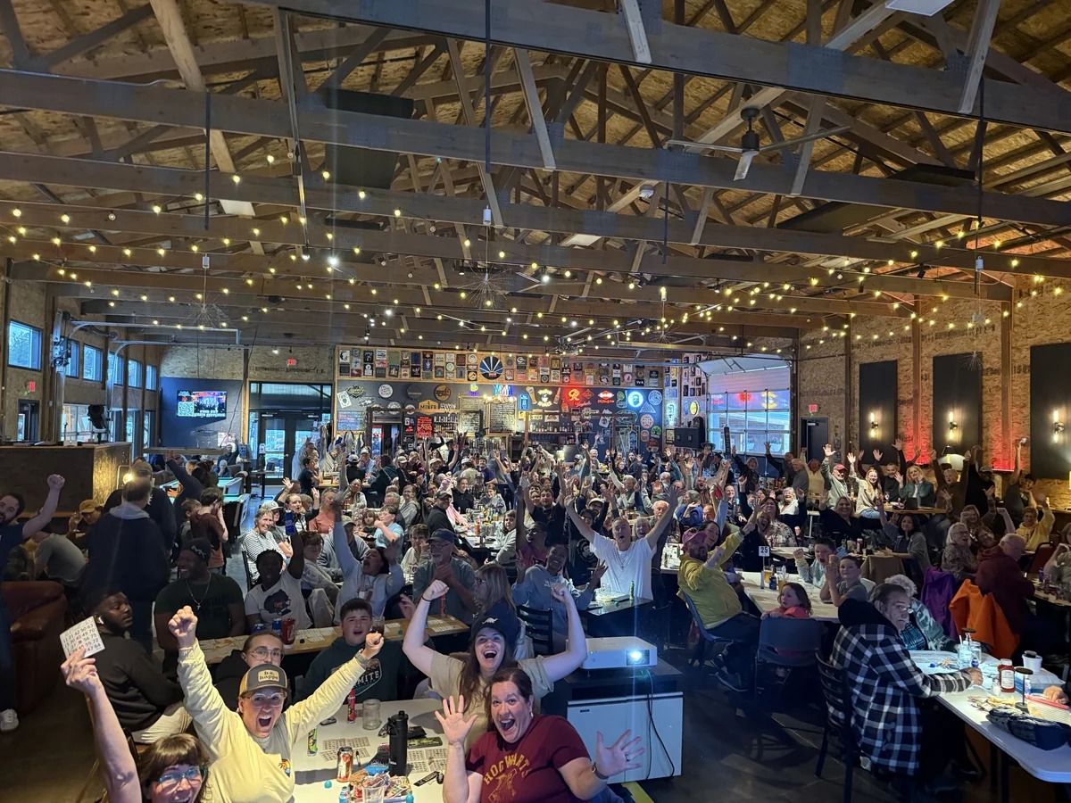 Bingo players at tables in the Custer Beacon raise their hands and bingo cards beneath exposed ceiling beams and string lights.